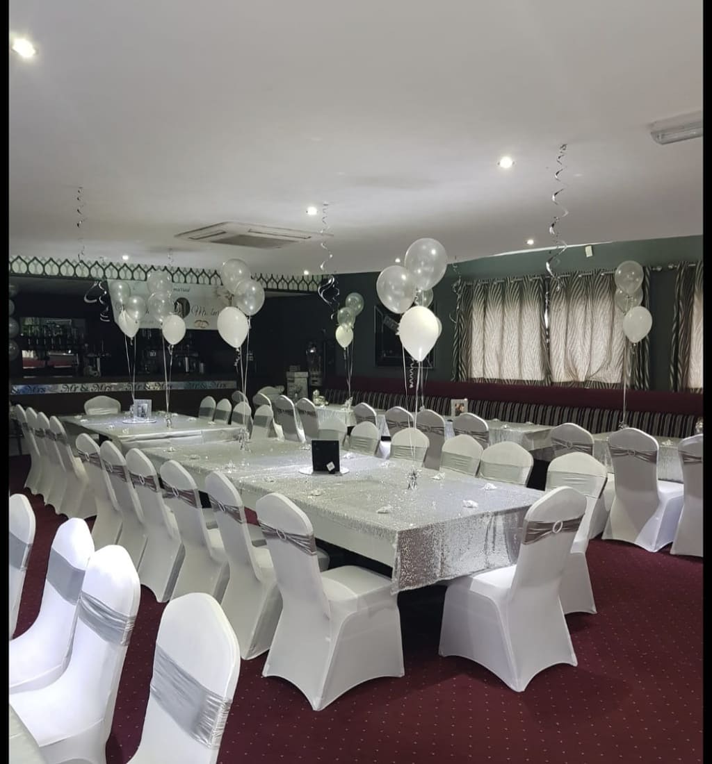 The function room laid out for a wedding, long tables in white cloths with covered chairs and silver balloons