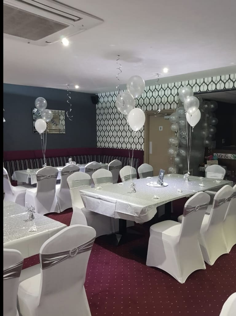 Square tables in silver cloths with covered chairs and balloons, set against the patterned wall of the function room